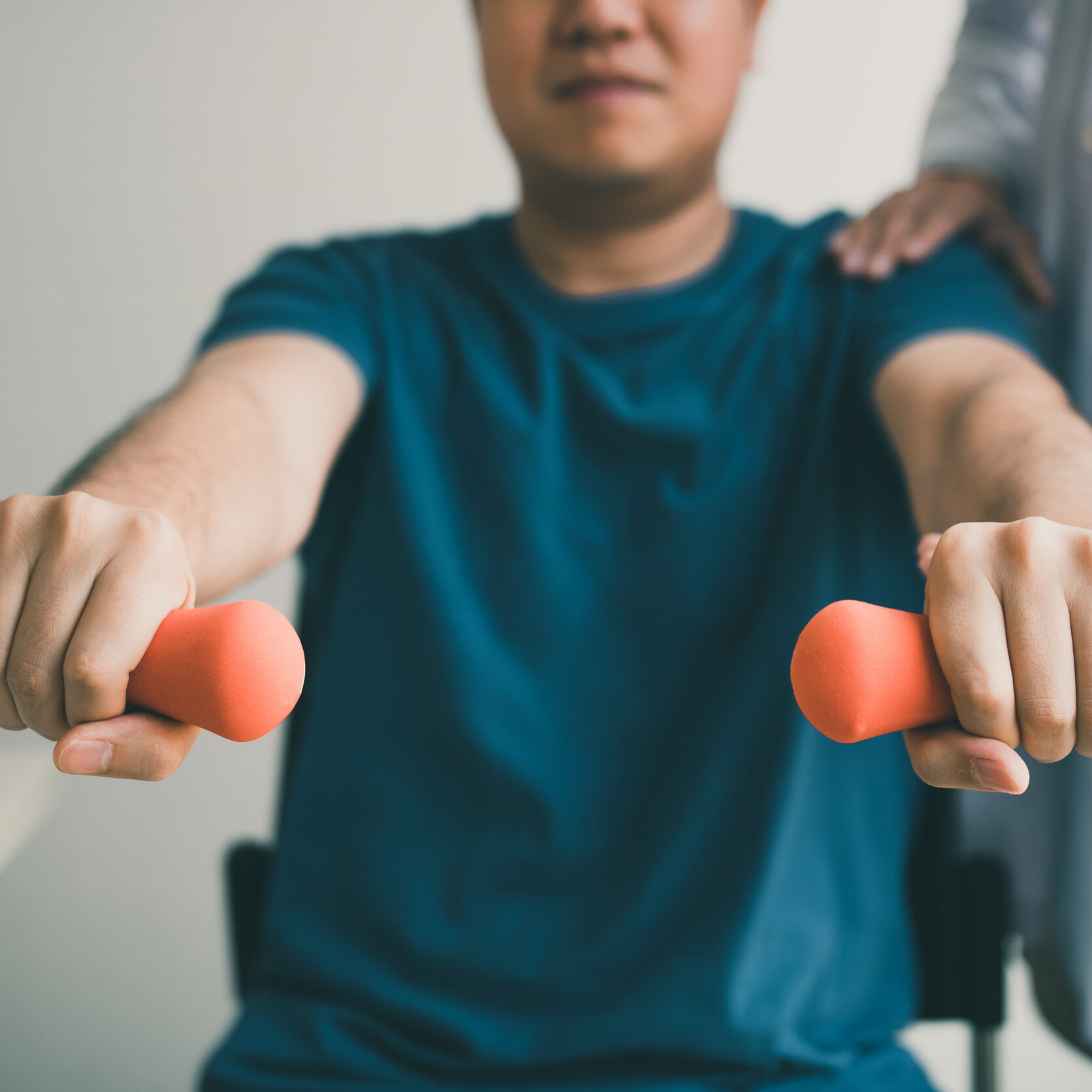 Asian young male physiotherapist helping patient with lifting dumbbells exercises in office.
